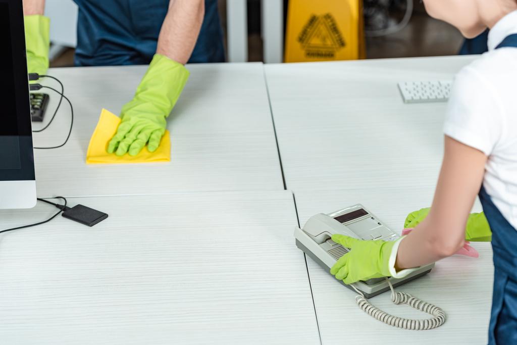 visão parcial do limpador limpando telefone do escritório perto da mesa de lavagem colega
 - Foto, Imagem