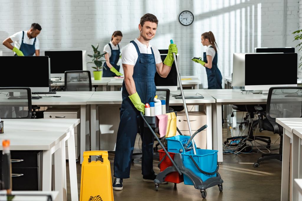 cheerful cleaner moving cart with cleaning supplies near multicultural colleagues - Photo, Image