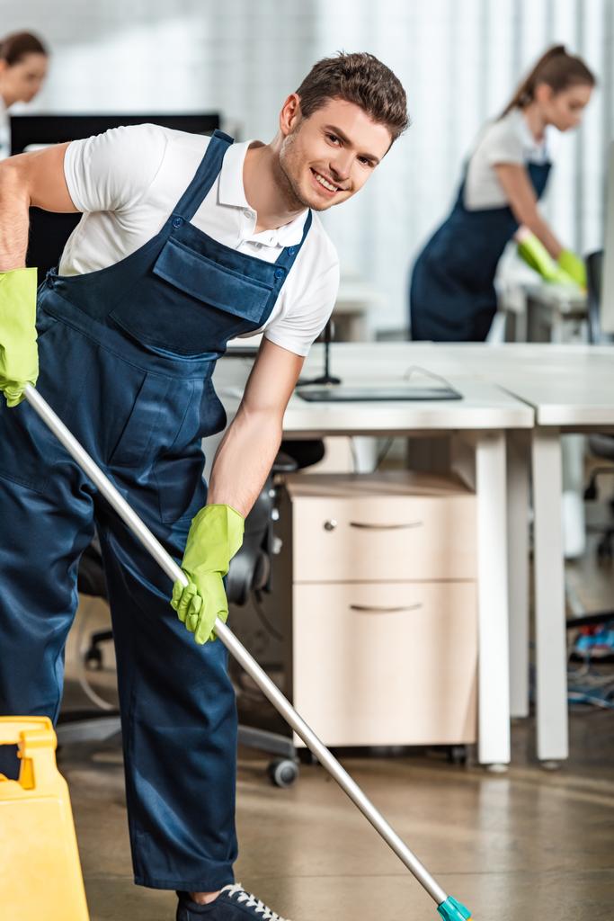 usmívající se čistič dívající se do kamery při mytí podlahy v kanceláři - Fotografie, Obrázek