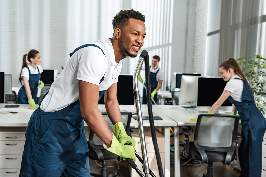 smiling african american cleaner moving vacuum cleaner near team of colleagues - Photo, Image