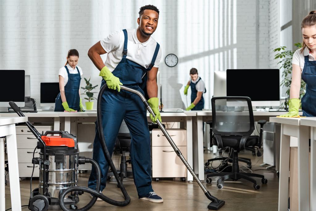 smiling african american cleaner vacuuming floor near team of multicultural colleagues - Photo, Image