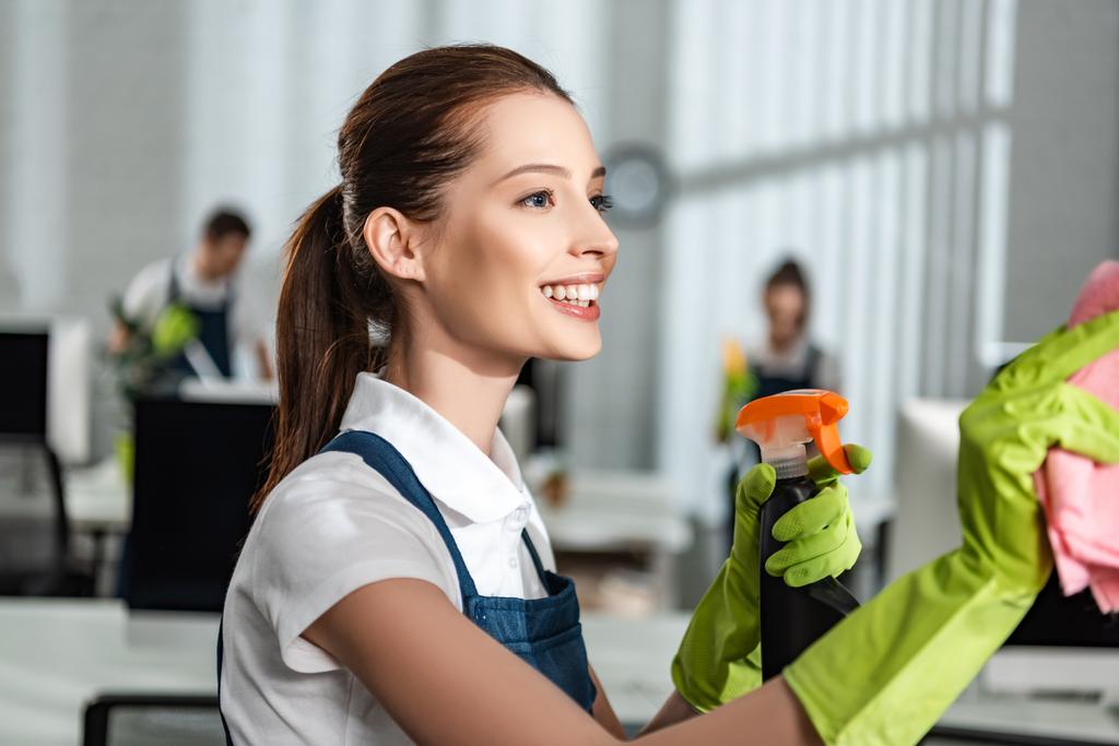 happy, attractive cleaner holding detergent spray and rag while cleaning office - Photo, Image