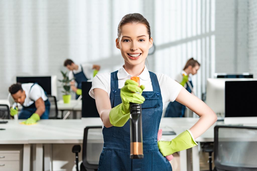 cheerful cleaner in overalls standing with hand on hip while holding spray bottle and looking at camera - Photo, Image