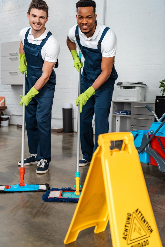 χαμογελαστοί πολυπολιτισμικοί καθαριστές που πλένουν το πάτωμα με σφουγγαρίστρες κοντά σε βρεγμένο δάπεδο - Φωτογραφία, εικόνα