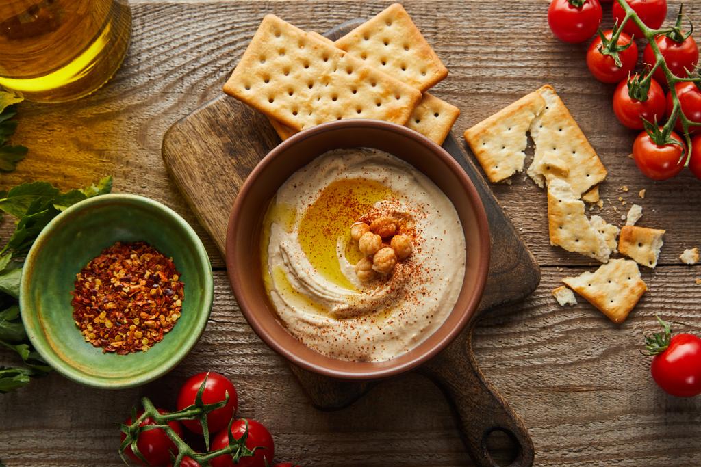 Top view of crackers, bowls with hummus and spices, vegetables, parsley and jar of olive oil on wooden background - Photo, Image