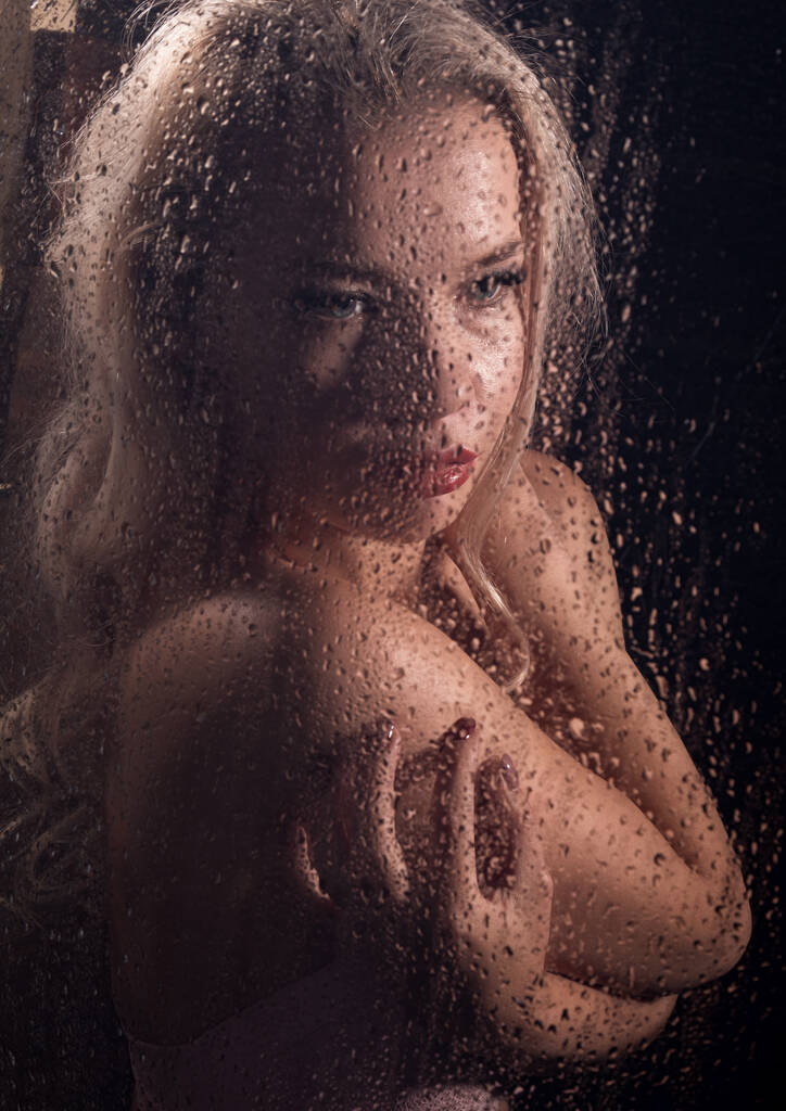 mooi blond vrouw achter nat glas op een donkere achtergrond, vrouw neemt een douche - Foto, afbeelding