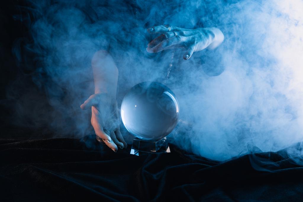 Vista recortada de bruja realizando ritual con bola de cristal sobre fondo azul oscuro
 - Foto, Imagen