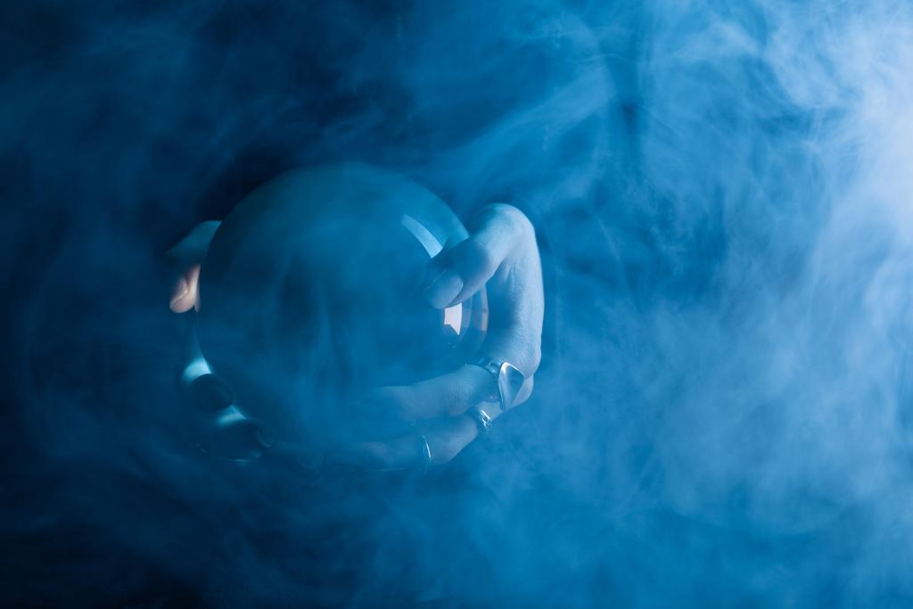 Oříznutý pohled na ženské ruce s křišťálovou koulí a kouř kolem na tmavomodrém pozadí - Fotografie, Obrázek
