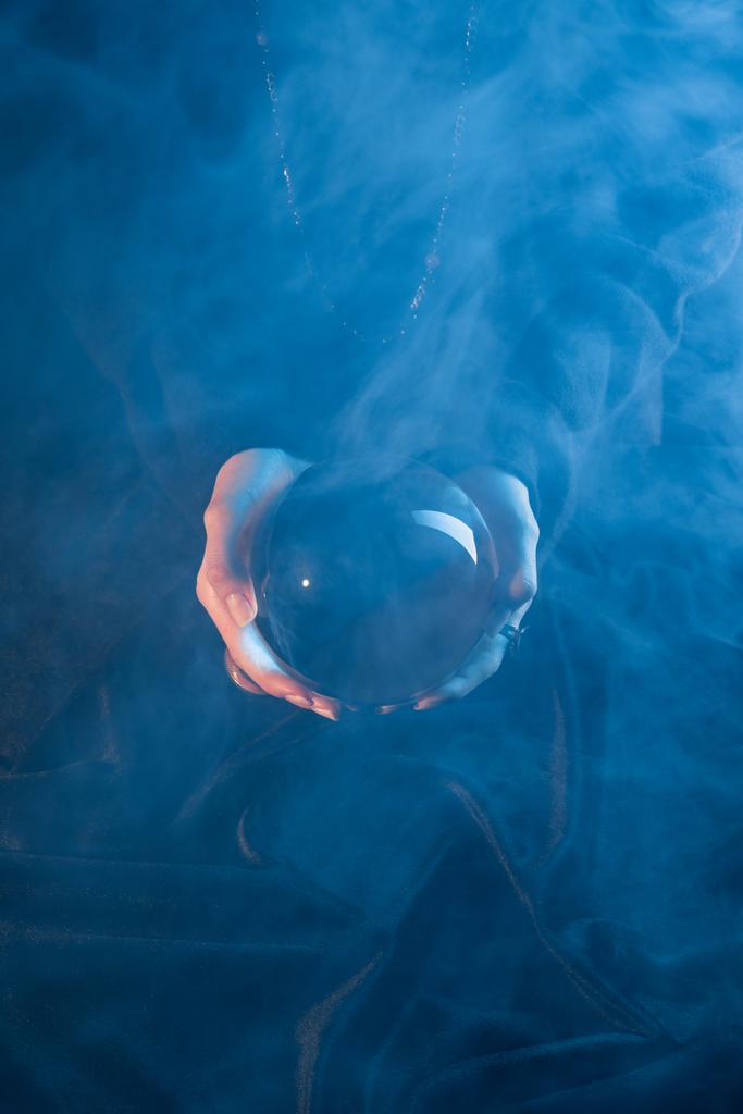 Cropped view of witch holding crystal ball above table on dark blue background - Photo, Image