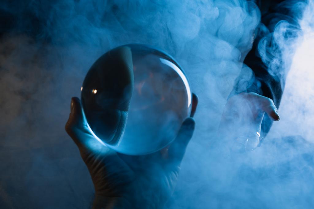 Cropped view of male hand with crystal ball and female hand with smoke around on dark blue - Photo, Image