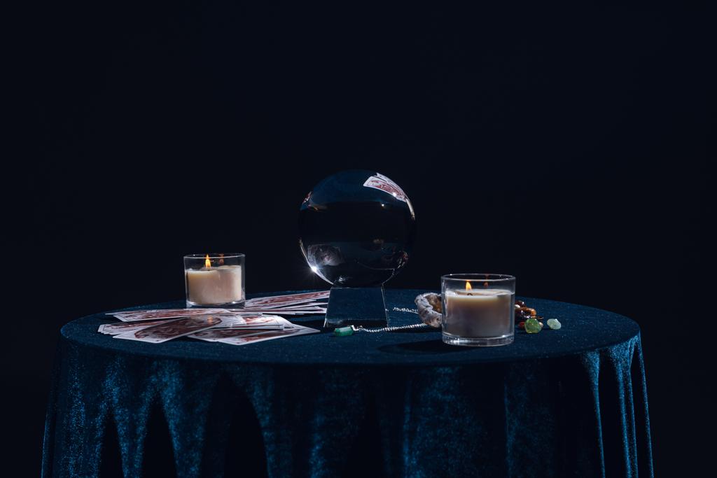Kiev, Oekraïne - 9 januari 2020: kristallen bol met kaarsen, tarotkaarten, stenen en ketting op ronde tafel geïsoleerd op zwart - Foto, afbeelding