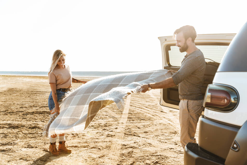 Foto van een positief jong geweldig liefdevol paar buitenshuis in de buurt van auto met mat afval op het strand. - Foto, afbeelding