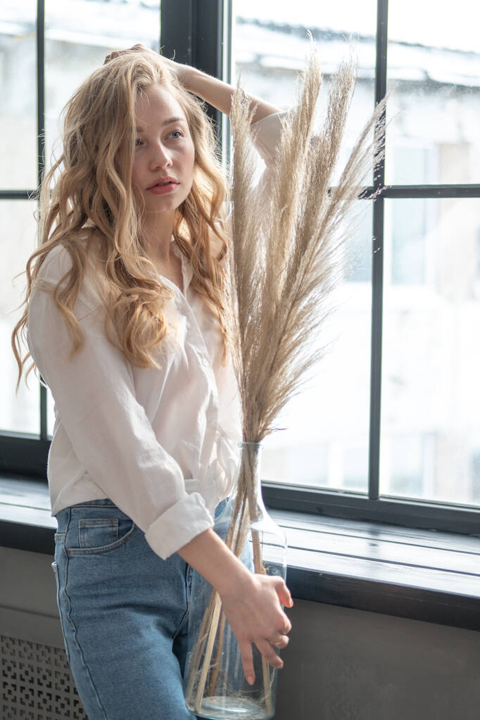 Junge hübsche blonde Frau in weißem Hemd und blauer Jeans, die neben dem Fenster steht. Morgenporträt eines Mädchens, das eine Vase mit trockenen Pflanzen hält. - Foto, Bild