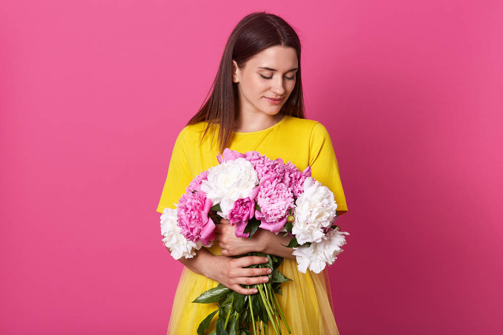 Horizontaal schot van zachte vrouw met roze en witte pioenroos bloemen, staat glimlachend isoalted over rooskleurige achtergrond, brunette dame draagt gele jurk, met cadeau voor 8 maart. Vrouwendag. - Foto, afbeelding