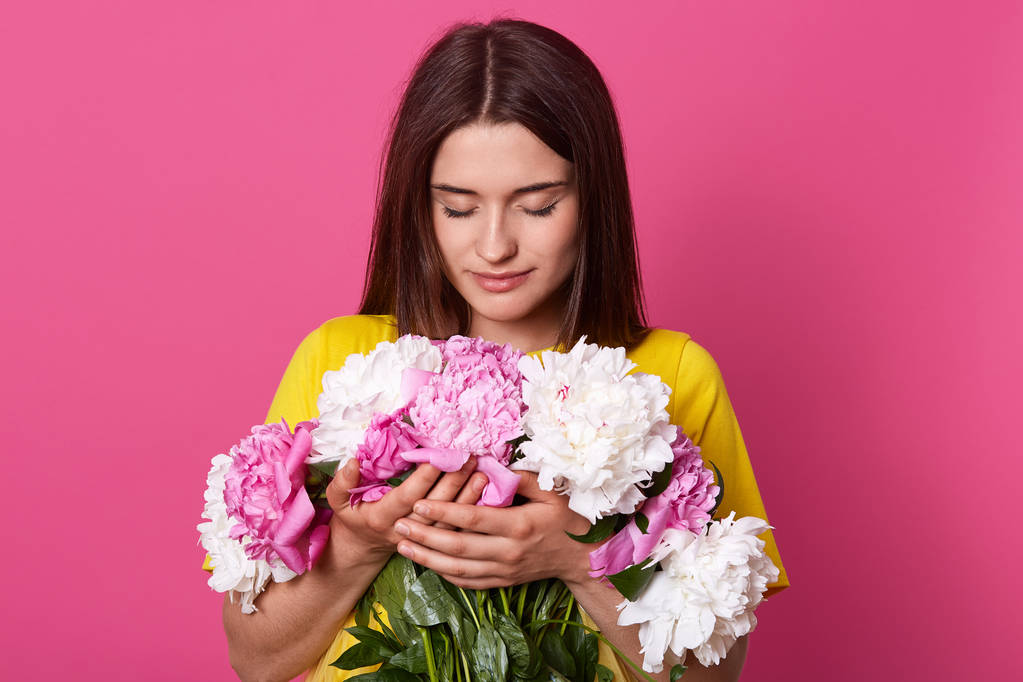 Plan intérieur de belle femelle aux cheveux foncés embrassant grand bouquet avec des fleurs de pivoine rose et blanc, a un regard doux, posant isolé sur fond de studio rose. Journée de la femme, 8 mars concept
. - Photo, image