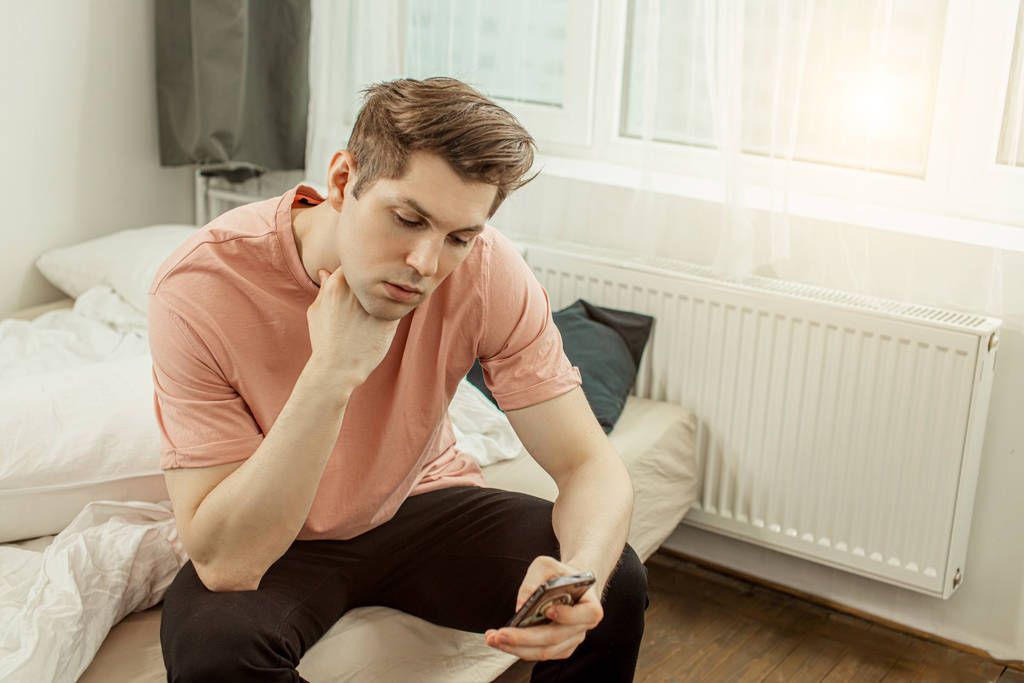 handsome caucasian young guy sitting on bed with smartphone - Foto, afbeelding