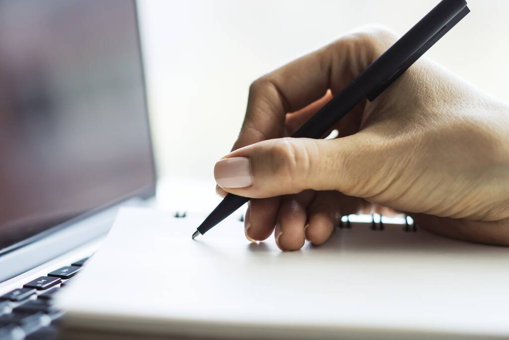 A menina escreve com uma caneta no bloco de notas no teclado do computador em um escritório ensolarado, conceito de negócios e educação. Fechar
 - Foto, Imagem