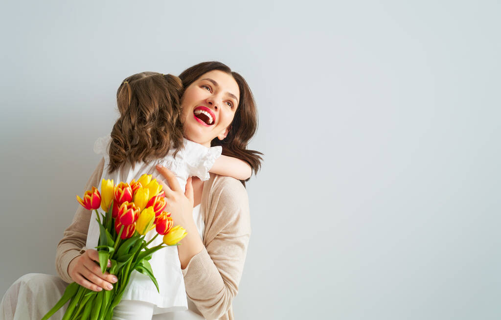 Счастливого женского дня! Ребенок поздравляет маму и дарит ей желтые цветы тюльпаны. Мама и девочка улыбаются на светло-сером фоне. Семейный праздник и единение
.  - Фото, изображение