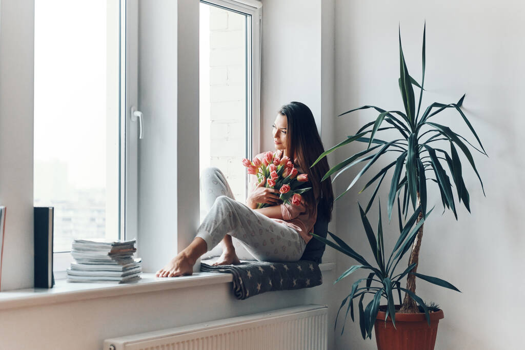 Houkutteleva nuori nainen kodikas pyjama nauttia hänen kimppu tulppaanit kun lepää ikkunalaudalla kotona
 - Valokuva, kuva