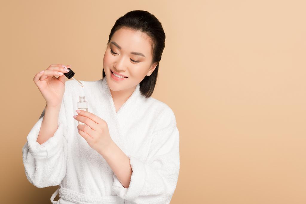 smiling beautiful asian woman in bathrobe holding dropper and bottle with serum on beige background - Photo, Image