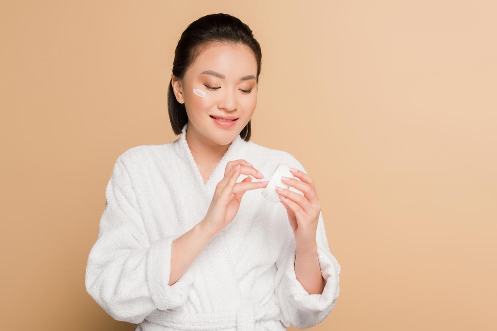 hymyilevä kaunis aasialainen nainen kylpytakissa soveltamalla kasvovoide beige tausta
 - Valokuva, kuva