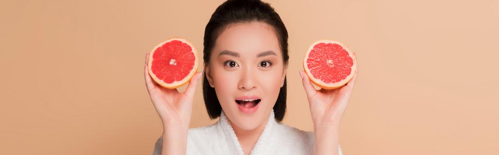 šokované krásné asijské žena v županu s grapefruit půlky izolované na béžové, panoramatický výstřel - Fotografie, Obrázek