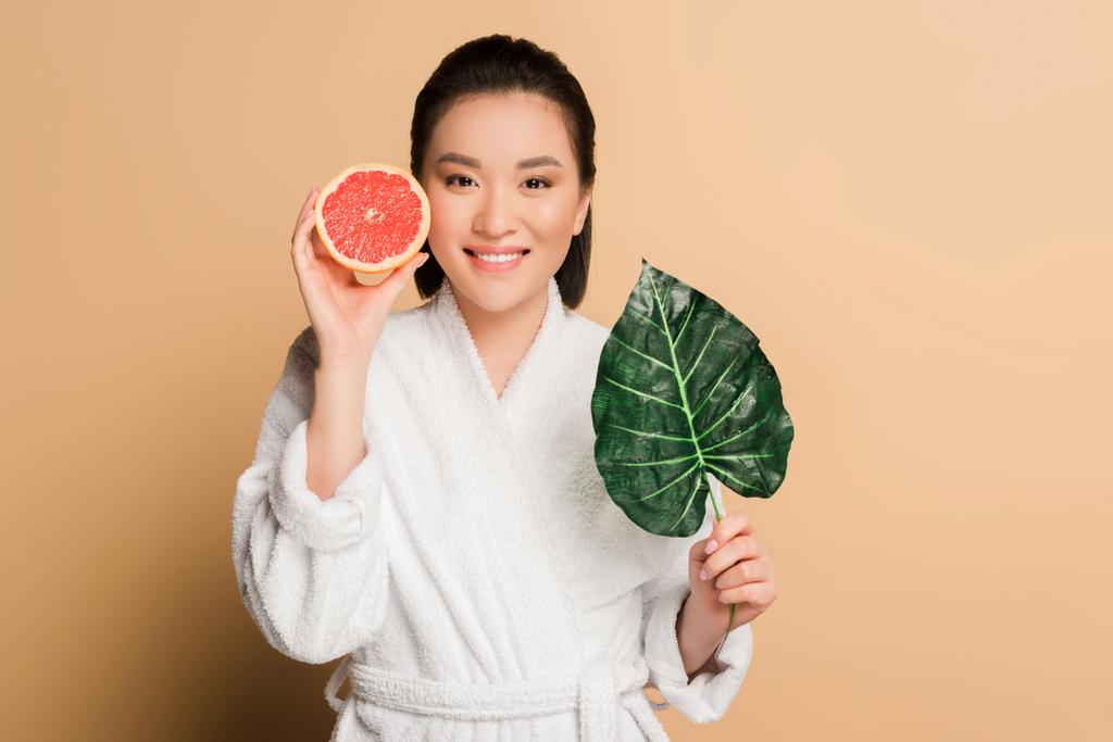 feliz hermosa mujer asiática en albornoz con pomelo mitad y hoja sobre fondo beige
 - Foto, imagen