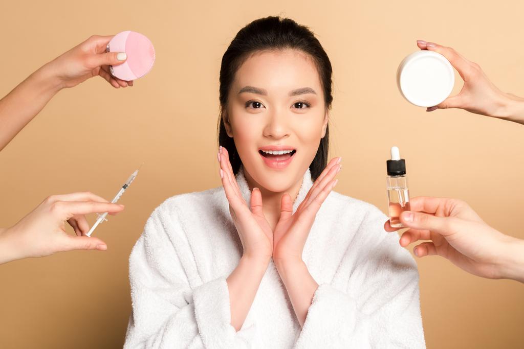 překvapená krásná asijská žena v županu v blízkosti rukou s obličejovým čisticím kartáčem, injekční stříkačkou, kosmetickým krémem a olejem na béžovém pozadí - Fotografie, Obrázek