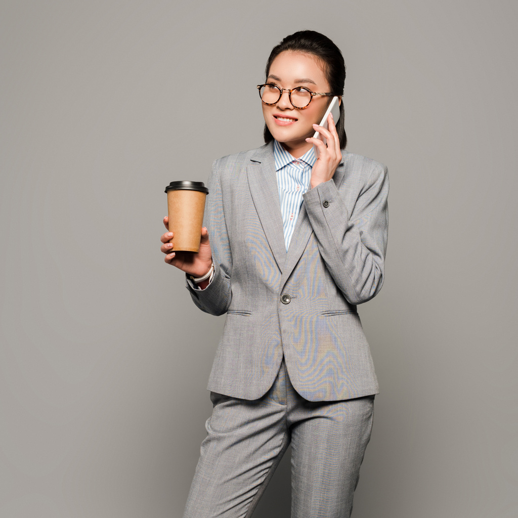 Lächelnde junge Geschäftsfrau mit Brille, Pappbecher in der Hand und Smartphone auf grauem Hintergrund - Foto, Bild