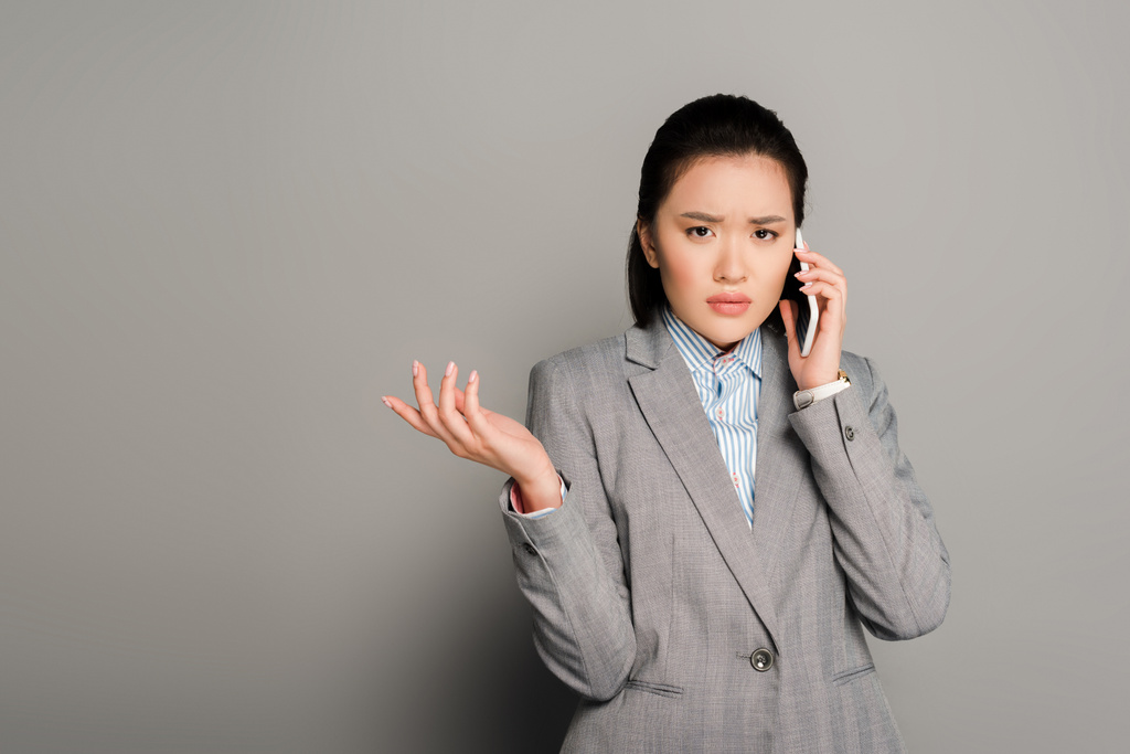confused young businesswoman in suit talking on smartphone on grey background - Photo, Image