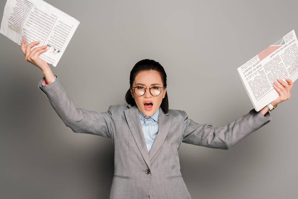 arrabbiato giovane donna d'affari in occhiali che tiene giornali su sfondo grigio
 - Foto, immagini