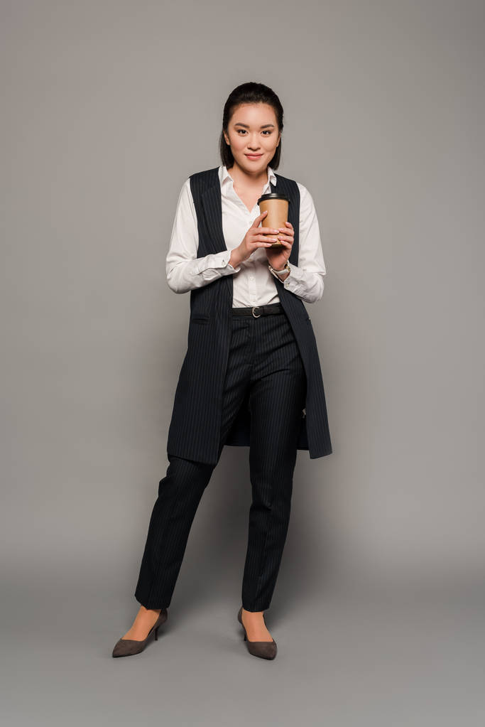 glimlachende jonge zakenvrouw houden koffie te gaan op grijze achtergrond - Foto, afbeelding
