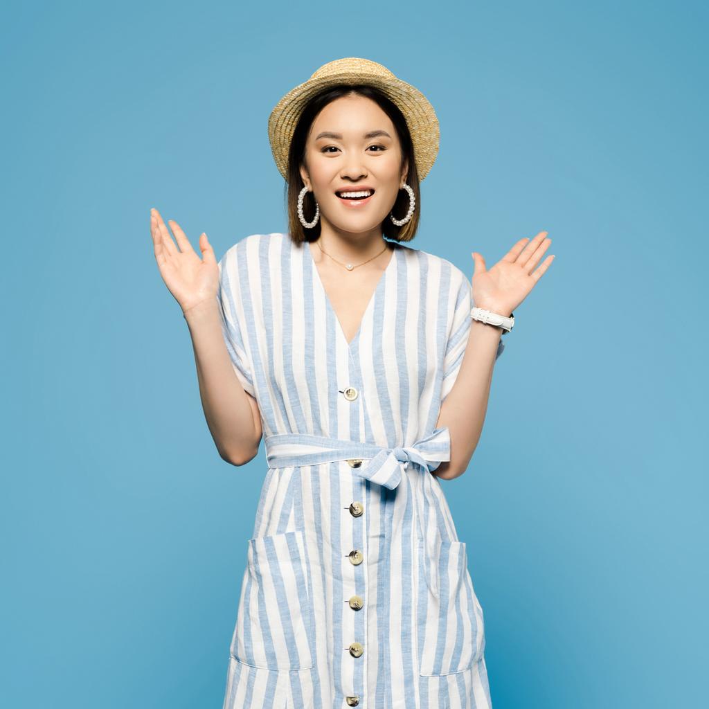 усміхнена брюнетка азіатська дівчина в смугастій сукні і солом'яному капелюсі з відкритими руками ізольована на синьому
 - Фото, зображення