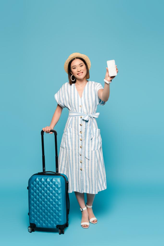 χαμογελαστή μελαχρινή Ασιάτισσα με ριγέ φόρεμα και ψάθινο καπέλο με τσάντα ταξιδίου και smartphone σε μπλε φόντο - Φωτογραφία, εικόνα