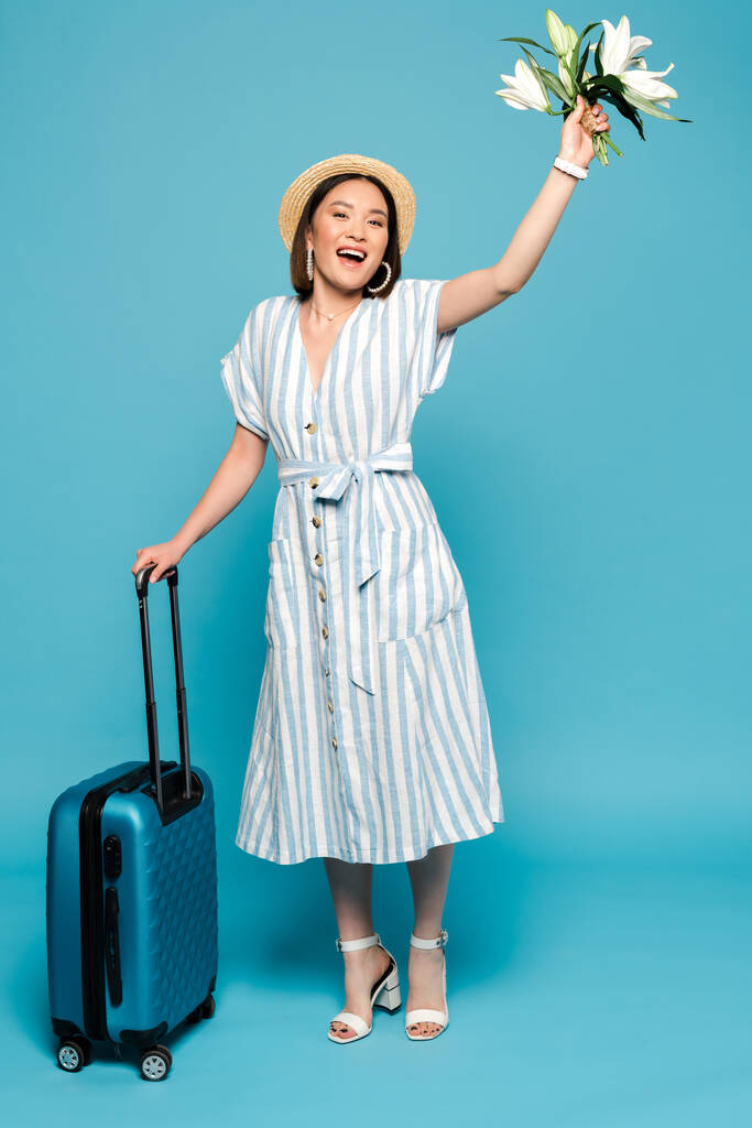 χαρούμενη μελαχρινή Ασιάτισσα με ριγέ φόρεμα και ψάθινο καπέλο με ταξιδιωτική τσάντα και μπουκέτο από κρίνα σε μπλε φόντο - Φωτογραφία, εικόνα
