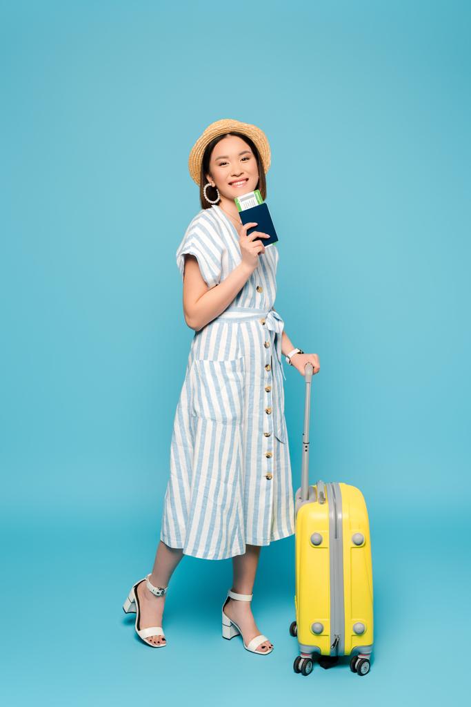 uśmiechnięta brunetka azjatycka dziewczyna w paski sukienka i słomka kapelusz z torba podróżna i paszport z biletem lotniczym na niebieskim tle - Zdjęcie, obraz