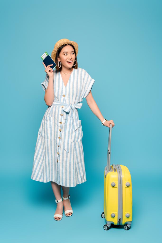 mosolygós barna lány csíkos ruha és szalma kalap utazási táska és útlevél repülőjegy kék háttér - Fotó, kép