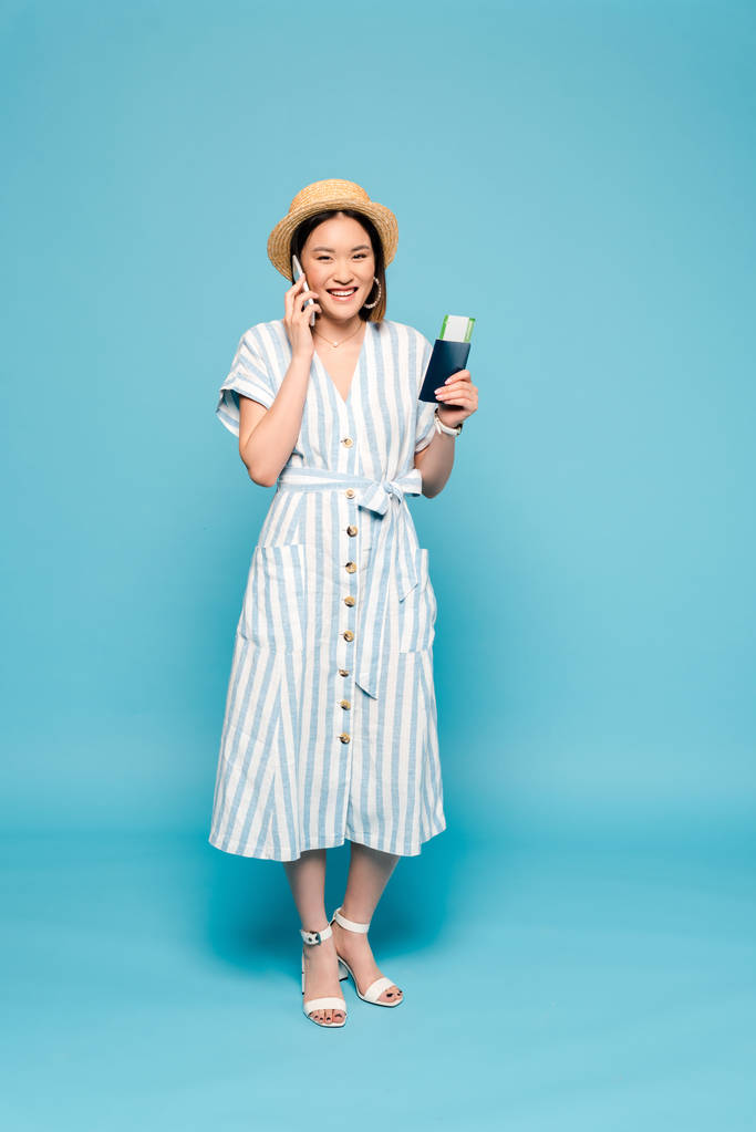 lächelnde brünette Asiatin in gestreiftem Kleid und Strohhut mit Pass und Flugticket im Gespräch auf Smartphone auf blauem Hintergrund - Foto, Bild
