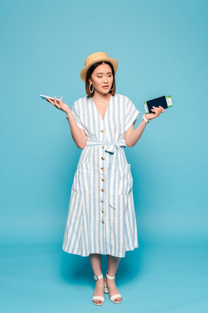 sad brunette asian girl in striped dress and straw hat with passport, air ticket, smartphone on blue background - Photo, Image