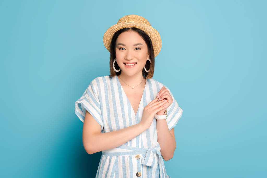 usmívající se brunetka asijské dívka v pruhovaných šatech a slamák klobouk na modrém pozadí - Fotografie, Obrázek