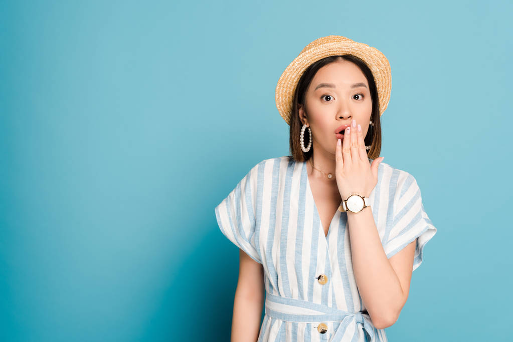 schockierte brünette asiatische Mädchen in gestreiftem Kleid und Strohhut bedeckt Mund mit Hand auf blauem Hintergrund - Foto, Bild