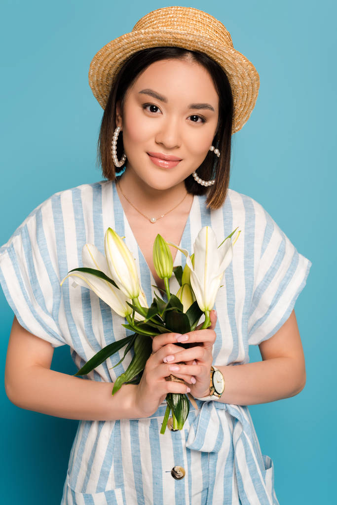 brunette asian girl in striped dress and straw hat holding bouquet of lilies on blue background - Photo, Image