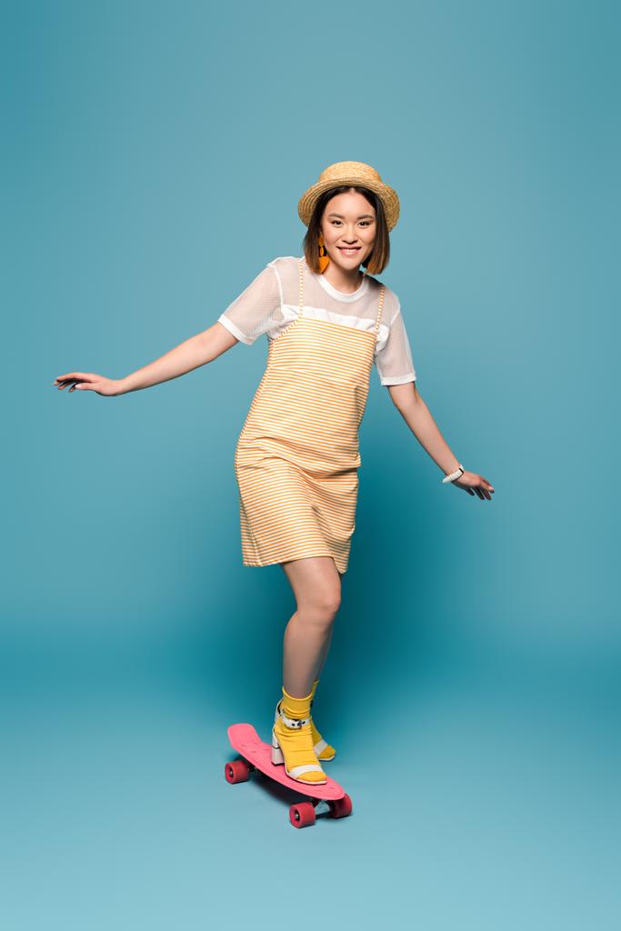 sonriente asiático chica en rayas amarillo vestido y paja sombrero cabalgando penny board en azul fondo
 - Foto, imagen