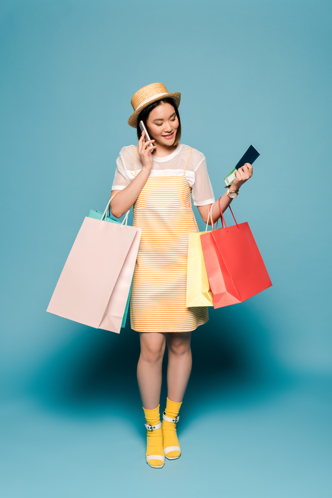 sonriente chica asiática en rayas vestido amarillo y sombrero de paja con bolsas de compras y pasaporte hablando en el teléfono inteligente sobre fondo azul
 - Foto, imagen