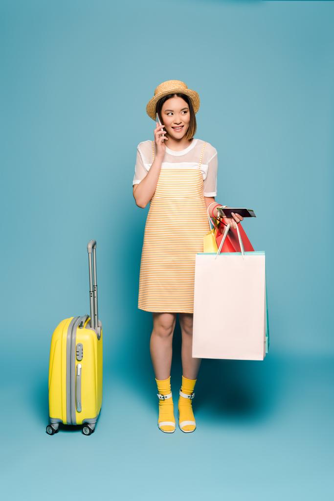 sonriente chica asiática en vestido amarillo rayado y sombrero de paja con bolsas de compras, pasaporte y maleta hablando en el teléfono inteligente sobre fondo azul
 - Foto, imagen