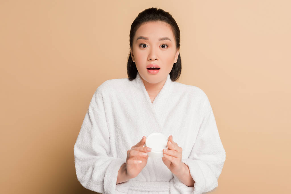 wstrząśnięta piękna azjatycka kobieta w szlafroku trzyma krem kosmetyczny na beżowym tle - Zdjęcie, obraz