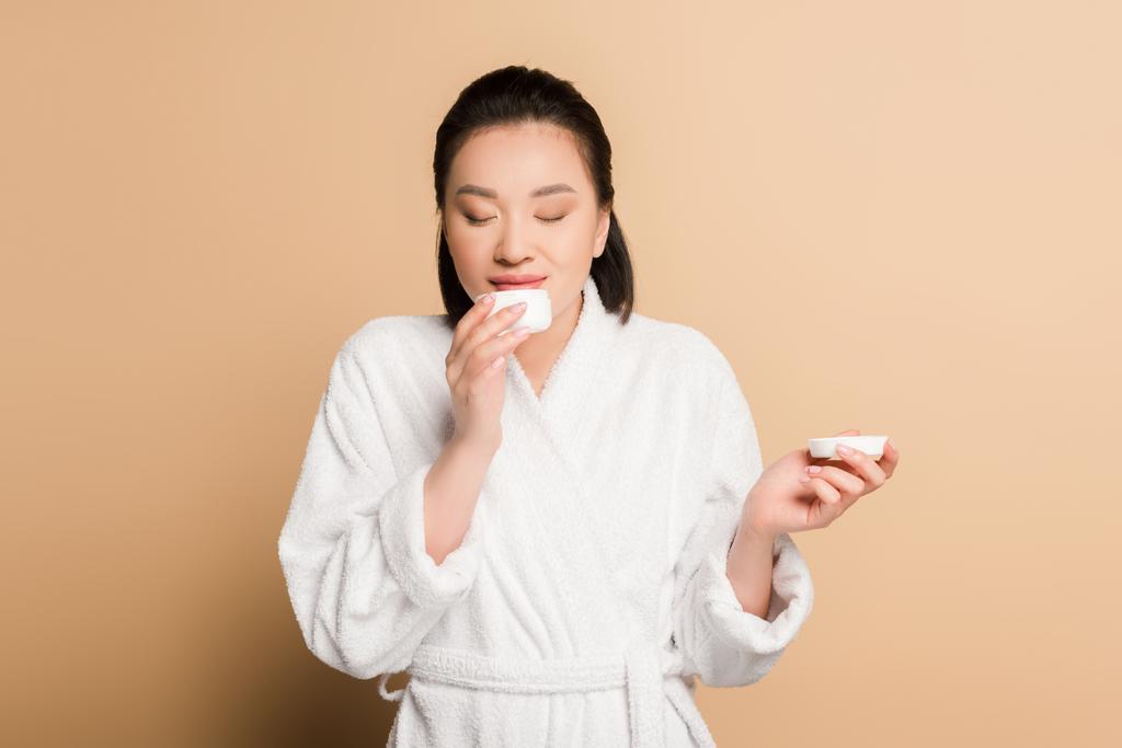 smiling beautiful asian woman in bathrobe smelling cosmetic cream on beige background - Photo, Image