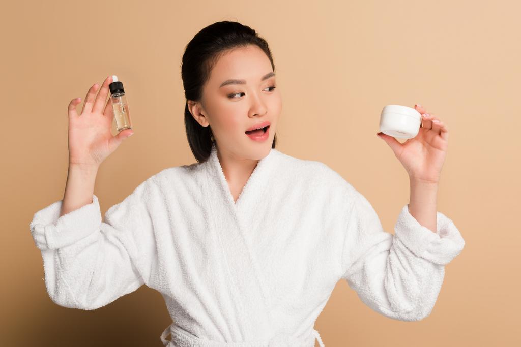 schöne asiatische Frau im Bademantel hält kosmetische Creme und Öl mit offenem Mund auf beigem Hintergrund - Foto, Bild
