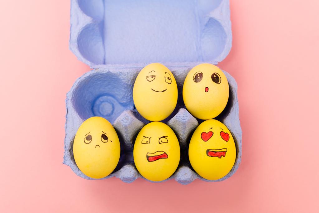 Κάτοψη των πολύχρωμων πασχαλινών αυγών με εκφράσεις του προσώπου στο δίσκο αυγών σε ροζ φόντο - Φωτογραφία, εικόνα
