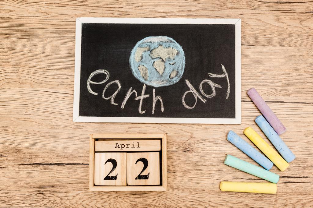 Top view of board with earth day lettering, pieces of chalk and calendar with 22 april inscription on wooden background - Photo, Image
